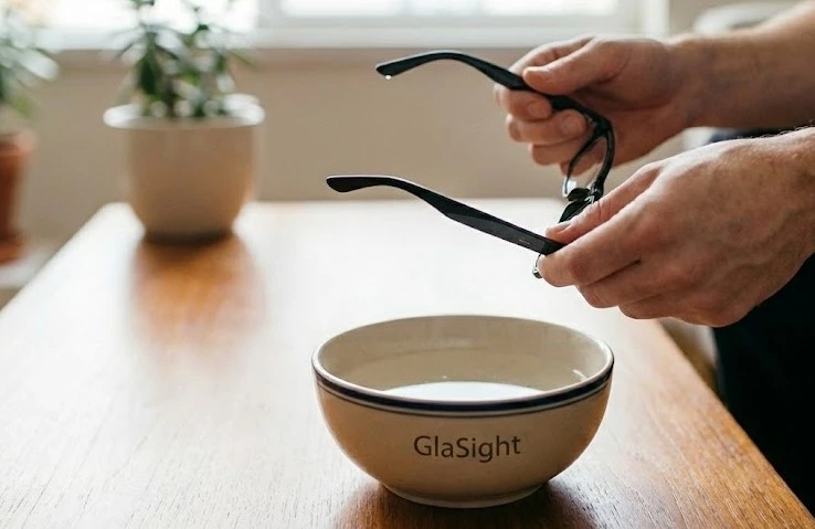Hands holding plastic eyeglass frames over a ceramic bowl of warm water to soften the material for safe bending and adjustment of glasses frame.