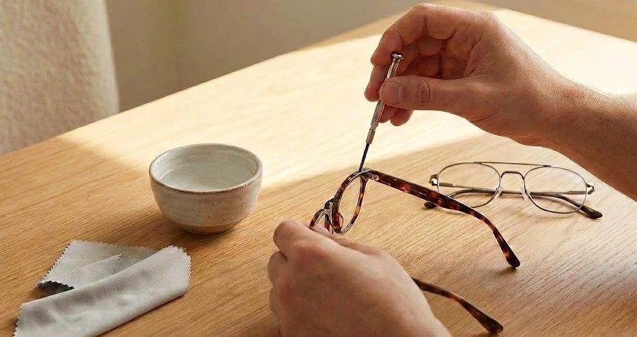 Close-up of hands tightening a loose hinge screw on eyeglasses using a micro-screwdriver, with a bowl of warm water and microfiber cloth on a wooden table for how to fix loose glasses DIY frame adjustment.