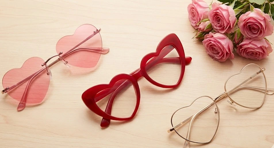 A comparison of the best styles of heart-shaped glasses for Valentine's Day 2026, featuring rimless pink frames, chunky red acetate frames, and gold wire heart frames arranged next to pink roses